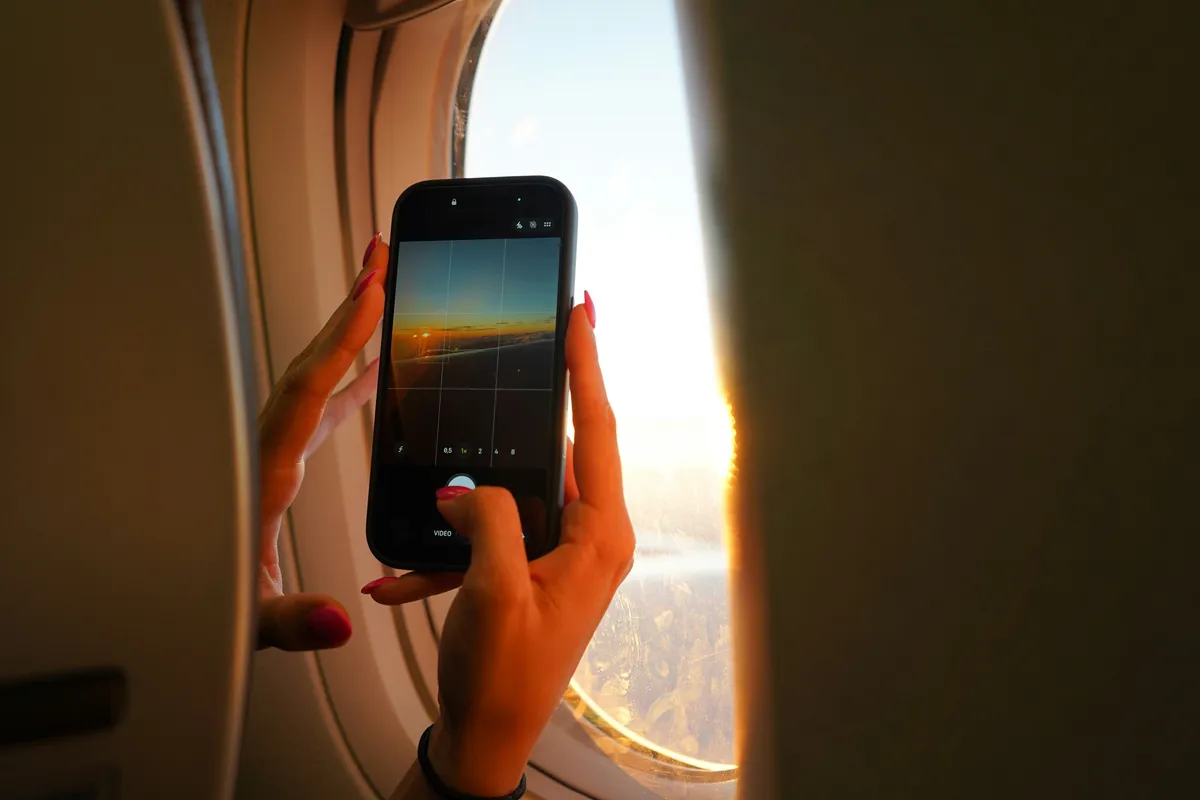 Uçak penceresinden gün batımını akıllı telefonla fotoğraflayan yolcunun elleri
